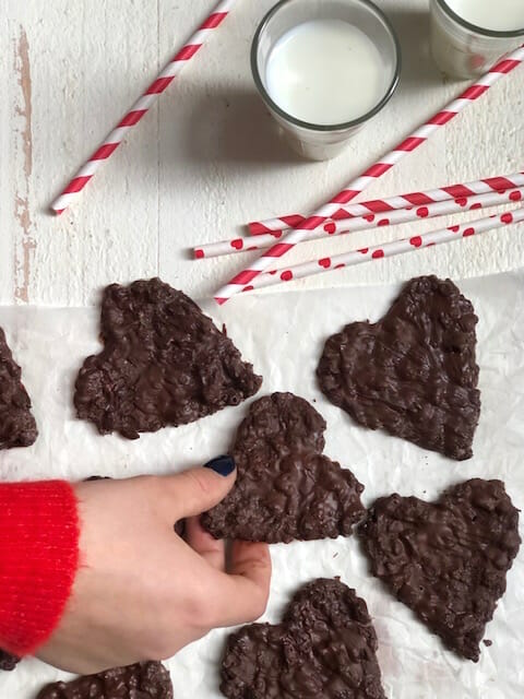 Heart shaped crispy chocolate coconut treats with glasses of milk, red and white straws, and a hand grabbing a treat