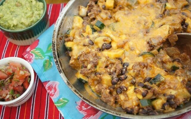 Mexican skillet supper of ground beef and beans topped with cheese in a skillet with colorful Mexican tablecloth and guacamole