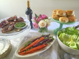 An Easter table set with lamb chops, carrots, biscuits, and other food with a white tablecloth