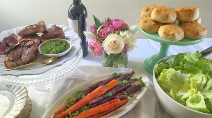 An Easter table set with lamb chops, carrots, biscuits, and other food with a white tablecloth