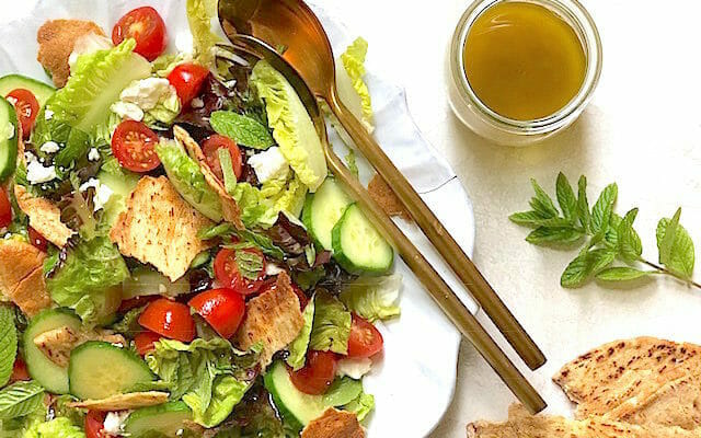 Fattoush salad on a white plate with gold salad servers and a jar of dressing