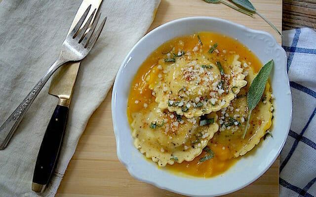 white plate with ravioli with butternut squash sauce and a napkin with fork and knife