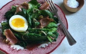 Warm spinach salad with soft egg in a vintage red bowl with fork and a bin of flaky salt
