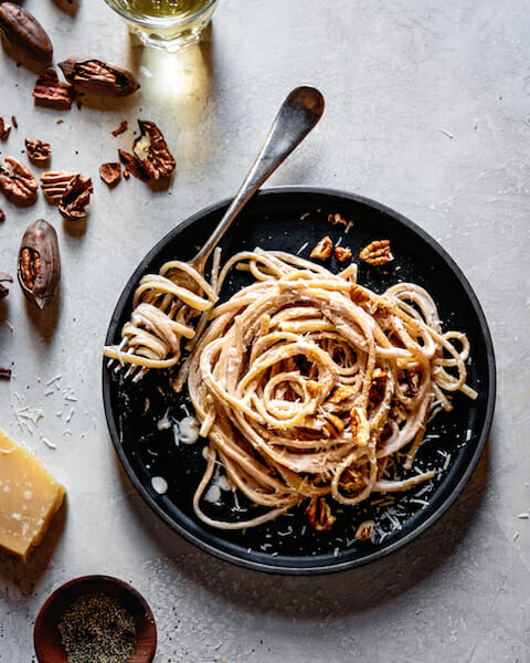 Linguine alfredo on a dark plate with a fork, parmesan, and pecans