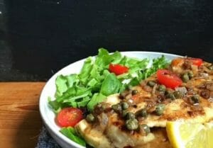 Chicken paillard with capers on a white plate with salad and tomatoes