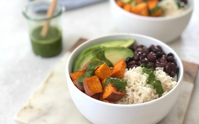 White bowl with sweet potato black beans avocado and a green sauce with a blue napkin