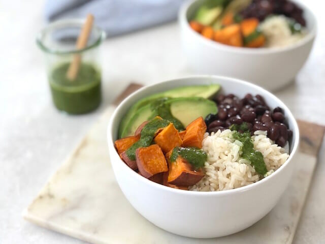 White bowl with sweet potato black beans avocado and a green sauce with a blue napkin