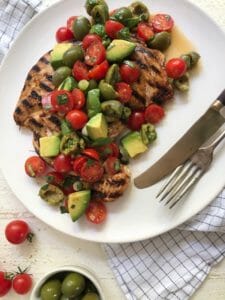 grilled chicken paillard on a white plate topped with tomato, olive, and avocado salad