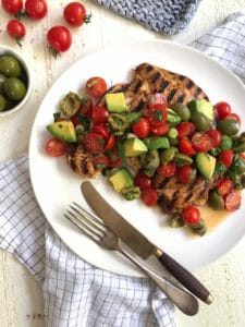 grilled chicken paillard with tomato, olive and avocado salad on a white plate with fork and knife