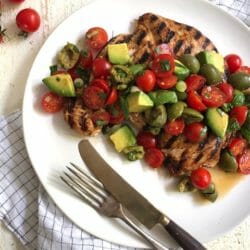 Grilled chicken paillard topped with tomato, olive, and avocado salad on a white plate with a fork and knife