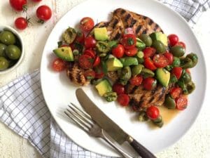 Grilled chicken paillard topped with tomato, olive, and avocado salad on a white plate with a fork and knife