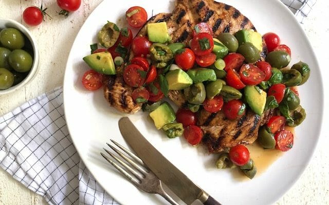 Grilled chicken paillard topped with tomato, olive, and avocado salad on a white plate with a fork and knife