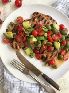 Grilled chicken paillard on a white plate topped with tomato, olive, and avocado salad